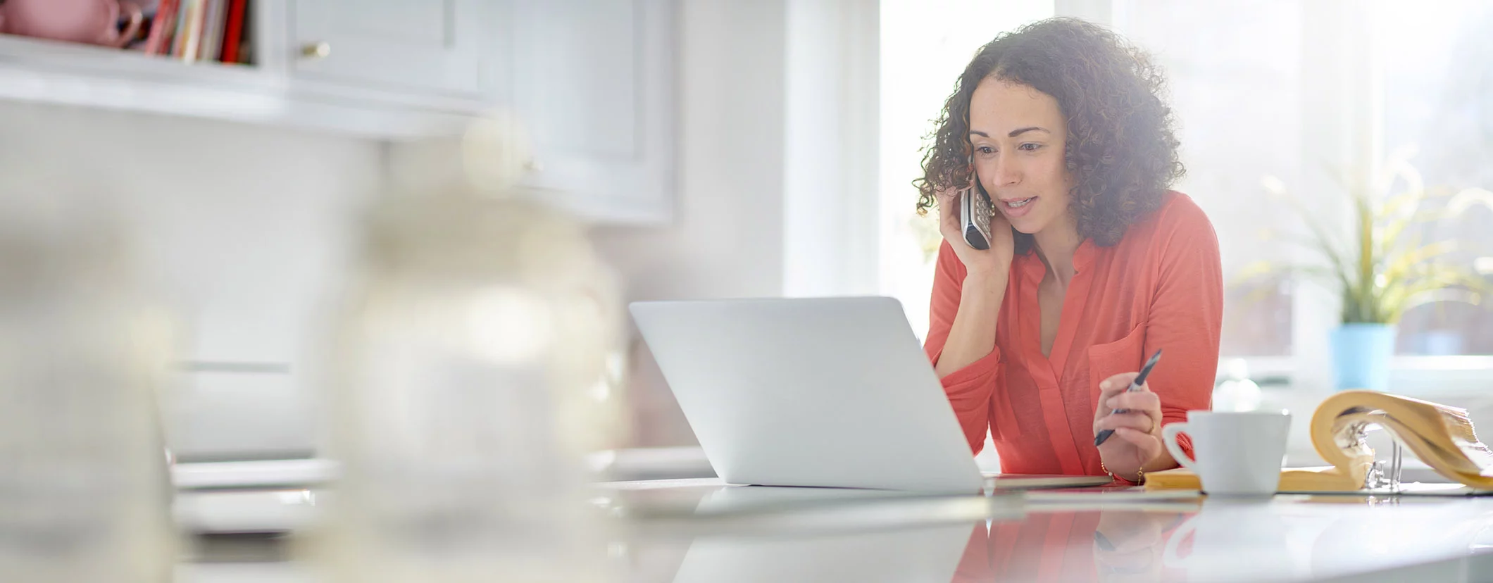 Professional remote executive assistant working from home, multitasking with a phone call, a laptop, and a notebook in a bright, organized kitchen setting.