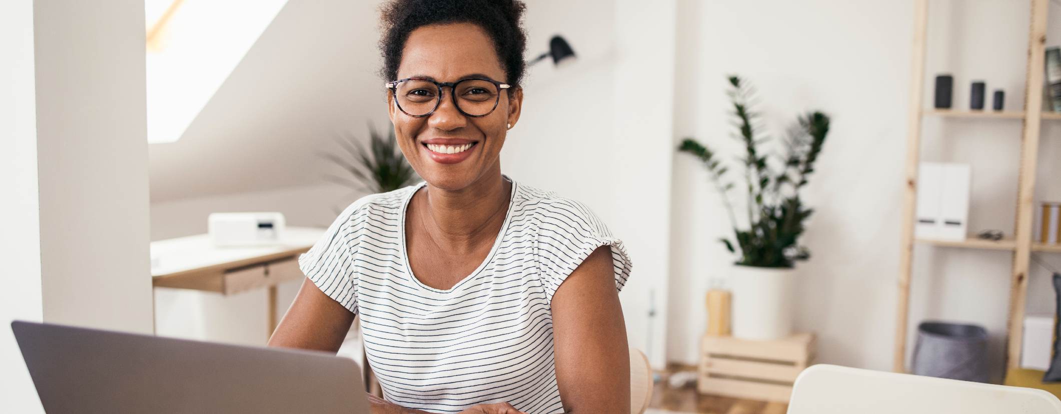 Woman sitting at laptop, smiling at camera in a brightly lit room