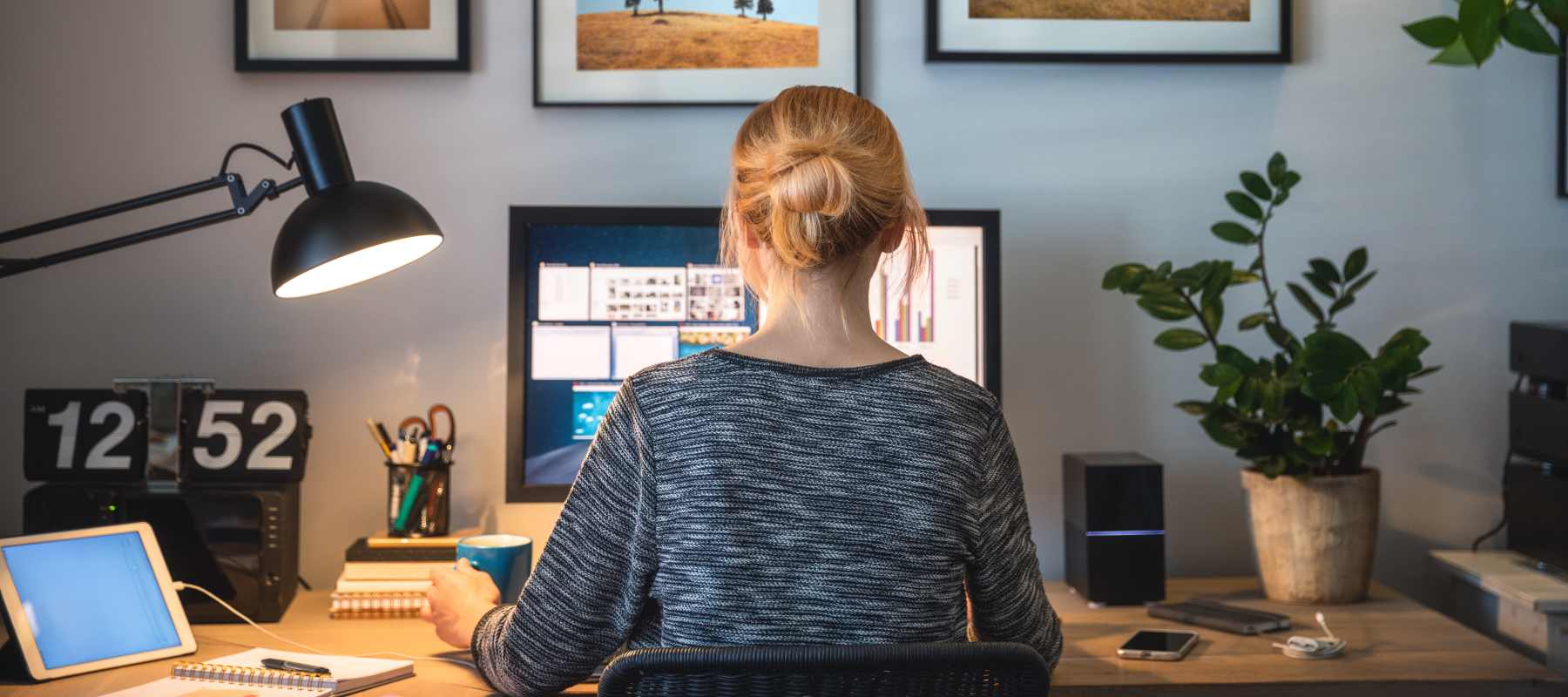 The image shows the back view of a woman sitting at a desk in a home office environment. She is wearing a gray sweater and has her hair pulled back. On the desk, there are various office supplies and equipment, including a computer monitor, a desk lamp, and a potted plant. The walls are decorated with framed artwork, and there is a clock visible on the wall.