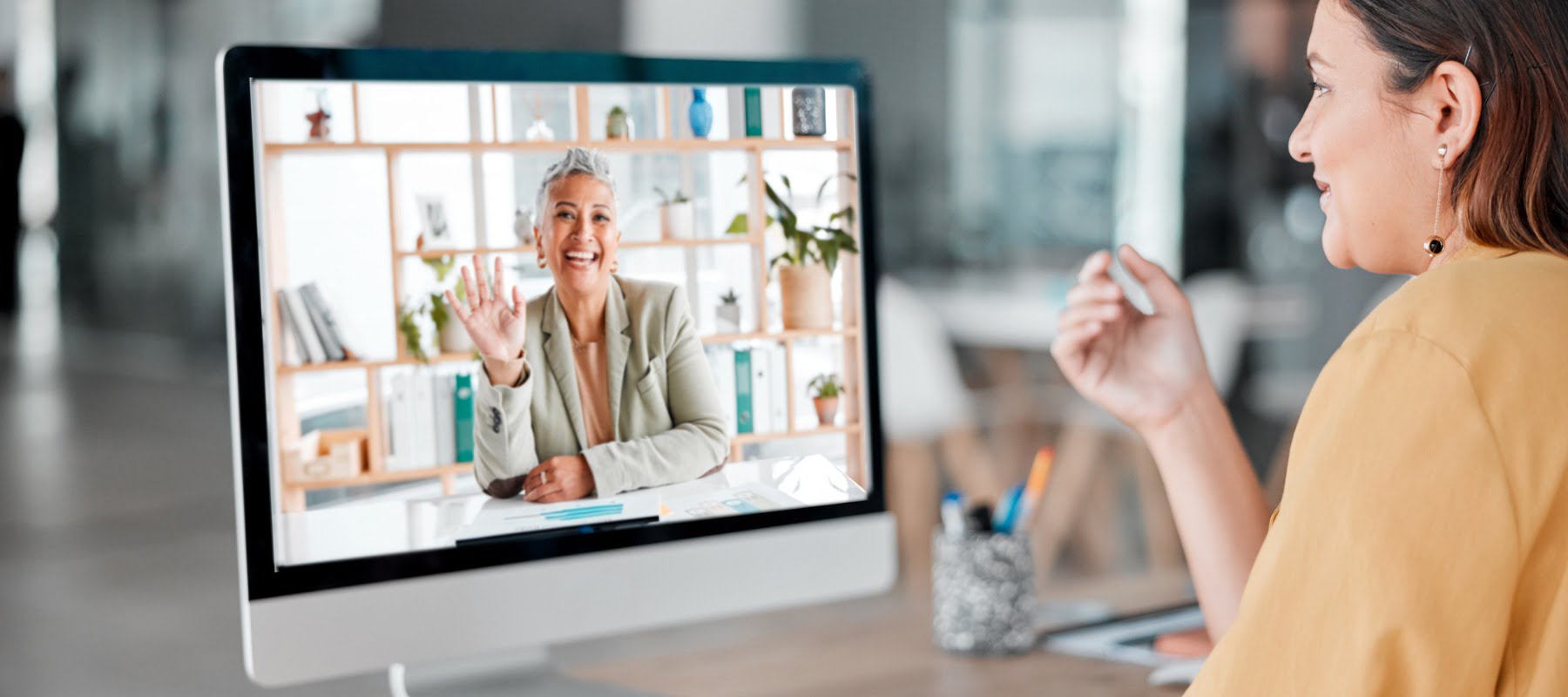 Woman remote executive assistant with short brown hair sitting at a desk looking at a computer monitor. She's talking to another woman via a video call. Both are looking engaged and productive.