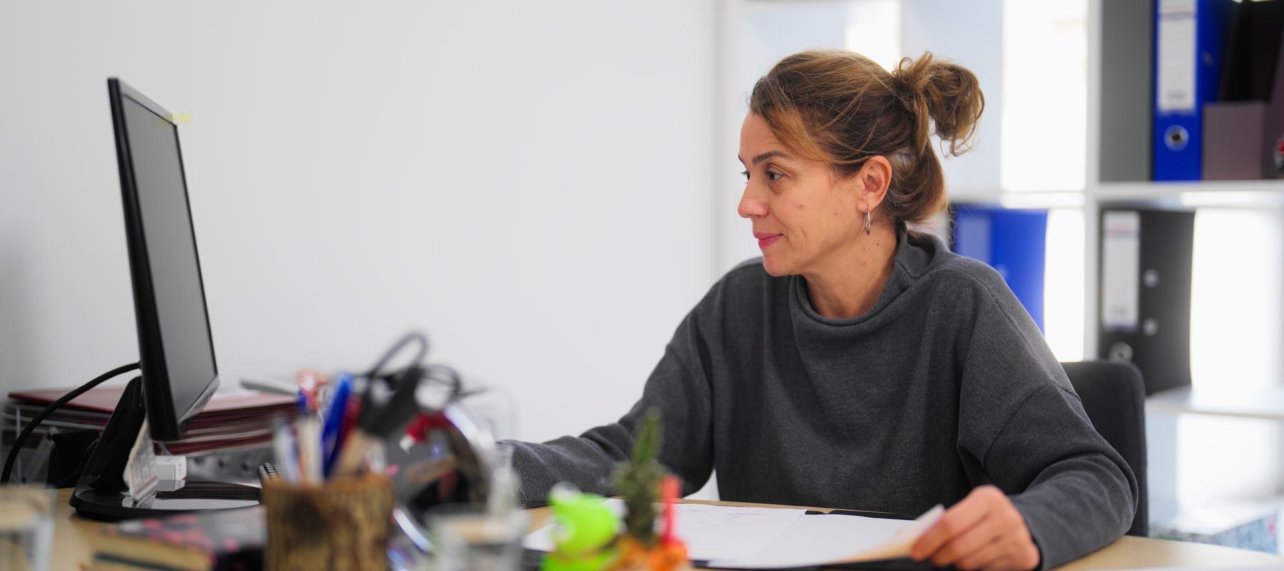 Woman with ponytail sitting at desk, looking at computer with papers in her hands.