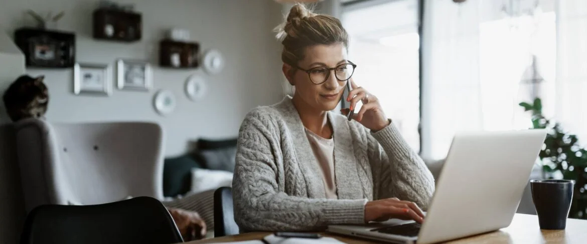 Executive assistant sitting at in-home office, working on laptop with phone.