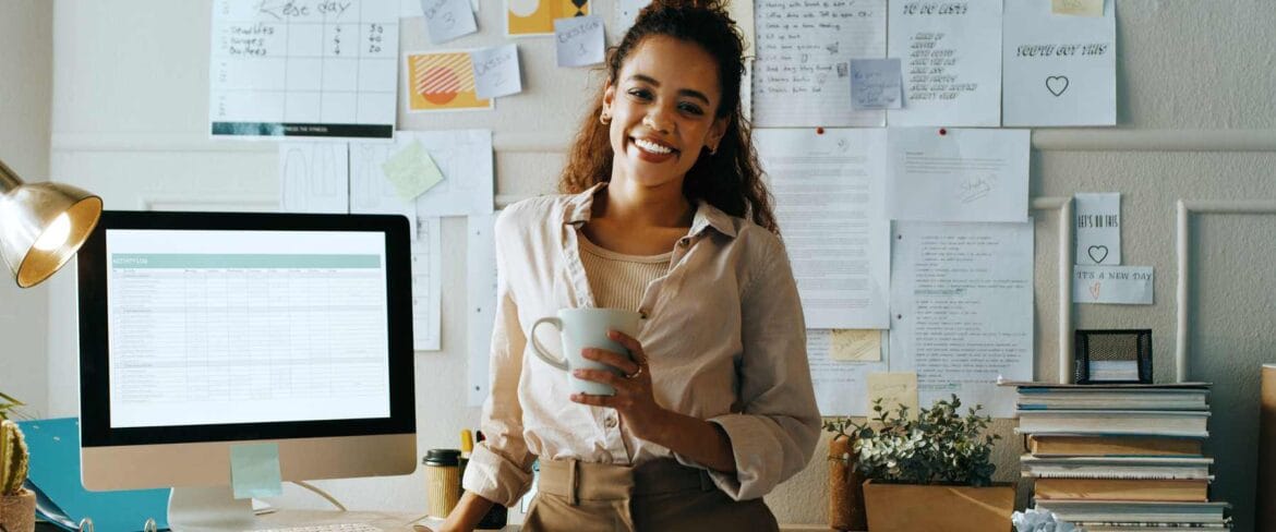 A smiling professional, exemplifying the virtual assistant vs. remote executive assistant comparison, stands at her organized workspace. She holds a coffee mug, surrounded by a desktop computer displaying a spreadsheet, stacks of books, and a wall covered in notes and schedules. Her casual yet professional attire and home office setup showcase the flexibility of remote work, while the organized environment highlights the skills needed for both virtual assistance and executive support roles.