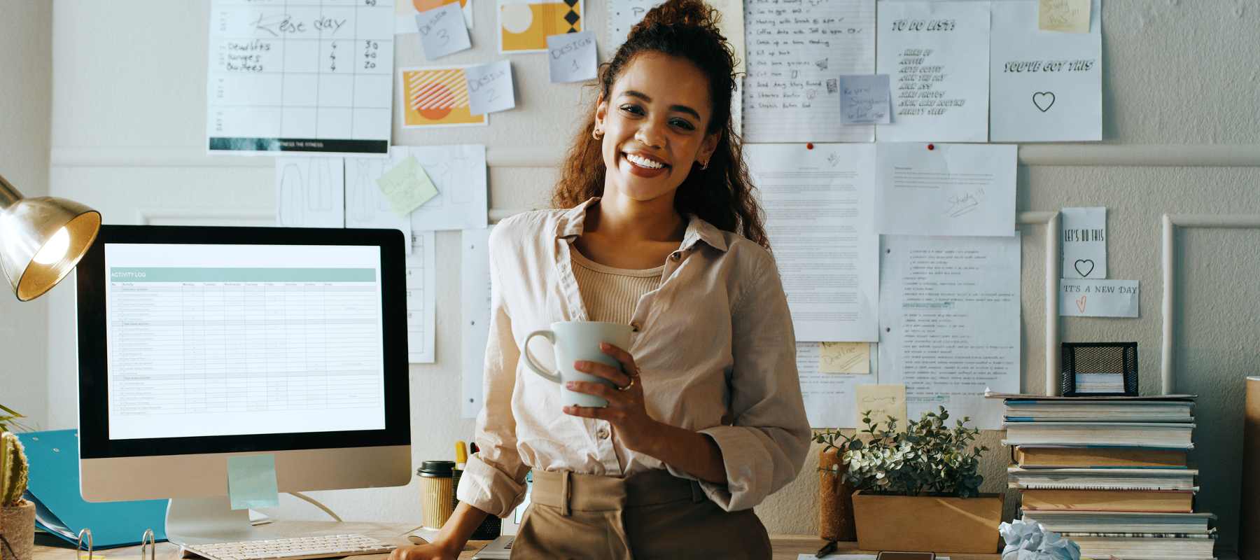 A smiling professional, exemplifying the virtual assistant vs. remote executive assistant comparison, stands at her organized workspace. She holds a coffee mug, surrounded by a desktop computer displaying a spreadsheet, stacks of books, and a wall covered in notes and schedules. Her casual yet professional attire and home office setup showcase the flexibility of remote work, while the organized environment highlights the skills needed for both virtual assistance and executive support roles.