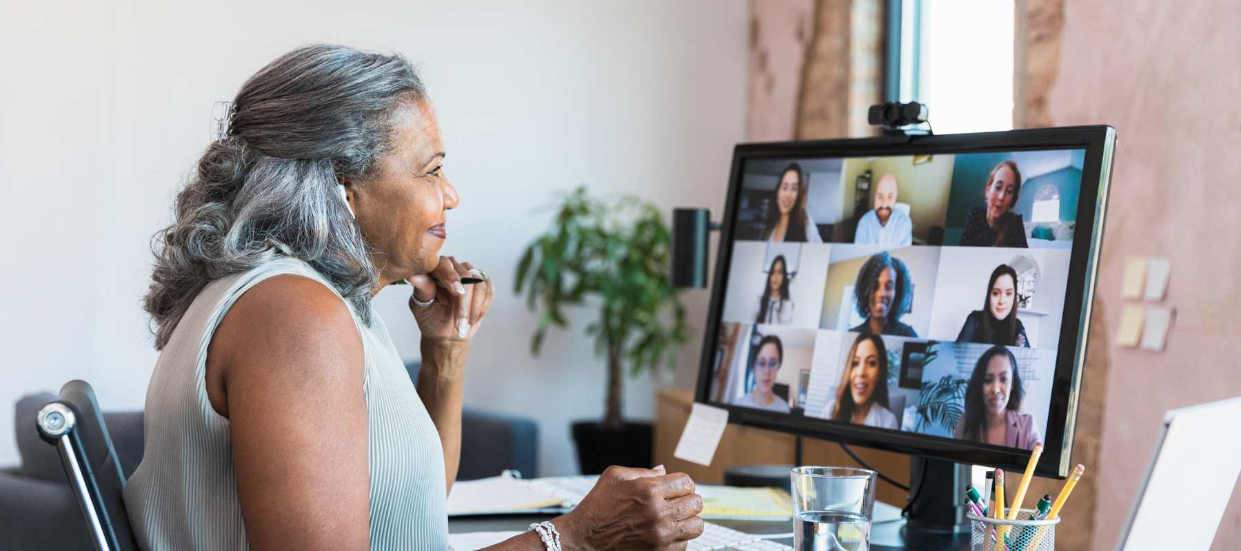 Woman managing a virtual team via video call on computer. She is older with grey hair, looking thoughtfully at the screen which shows different virtual employees.