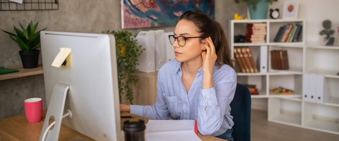 Young professional woman working as an executive assistant from a home office, focused on her desktop computer. The modern workspace includes a coffee mug, smartphone, and paperwork, with a bookshelf and abstract art in the background. Remote work, home office setup, and productivity.