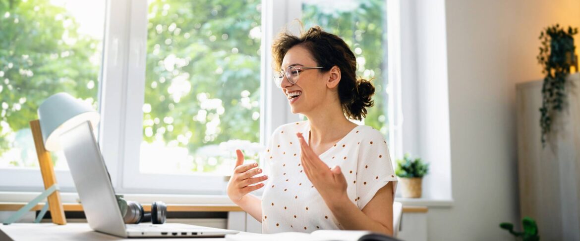 A young woman with short hair and glasses, wearing a white top with red polka dots, smiles while speaking in a bright home office. The room has large windows with a view of green trees, and her desk is equipped with a laptop, a desk lamp, and some plants.