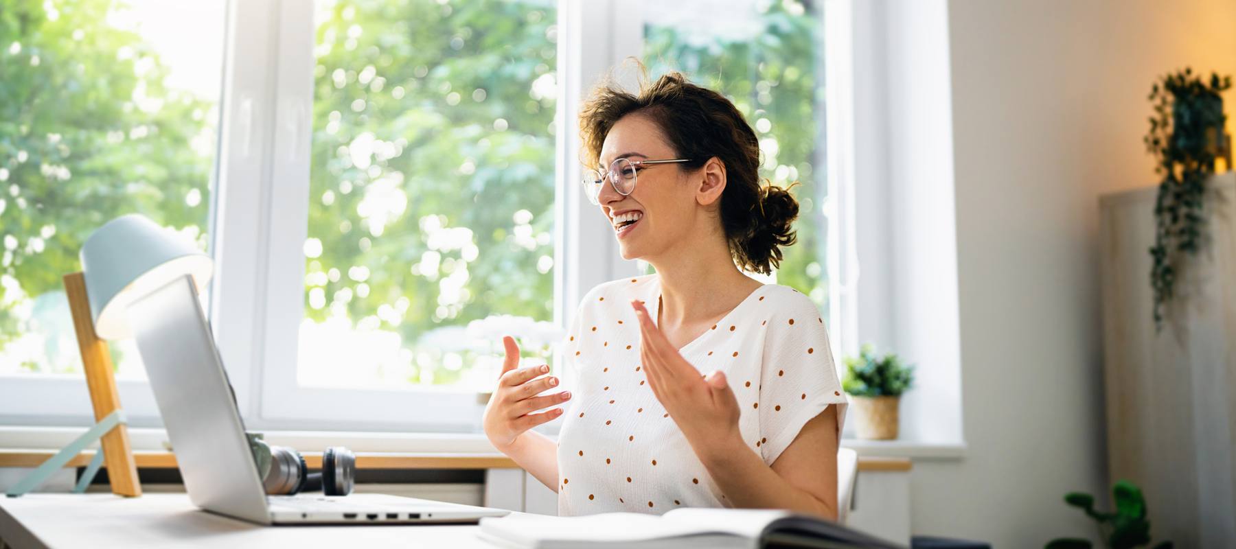 A young woman with short hair and glasses, wearing a white top with red polka dots, smiles while speaking in a bright home office. The room has large windows with a view of green trees, and her desk is equipped with a laptop, a desk lamp, and some plants.