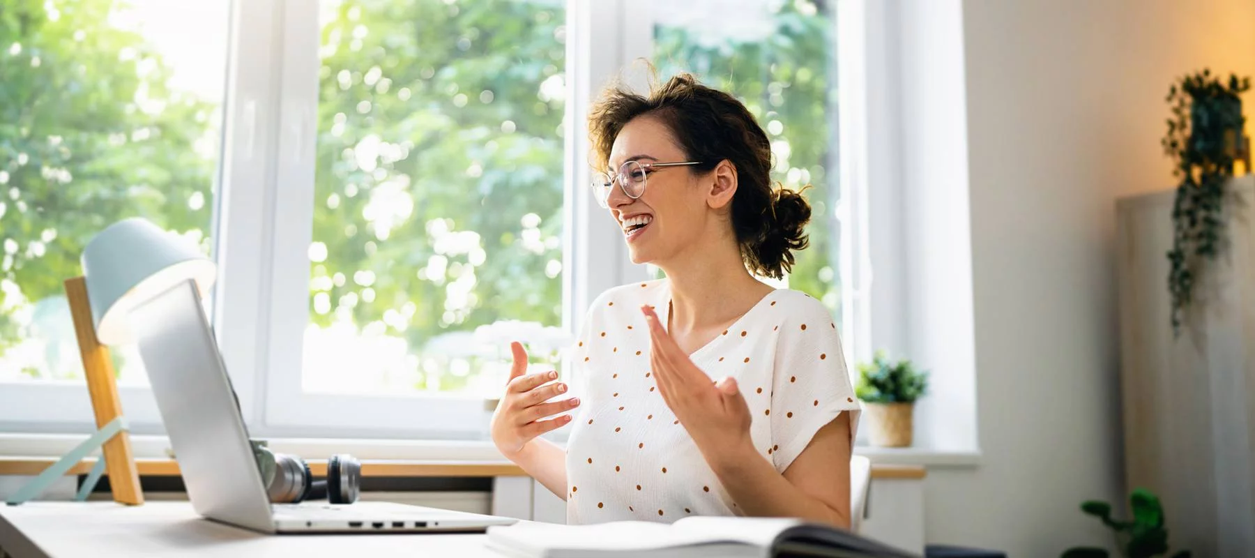 A young woman with short hair and glasses, wearing a white top with red polka dots, smiles while speaking in a bright home office. The room has large windows with a view of green trees, and her desk is equipped with a laptop, a desk lamp, and some plants.