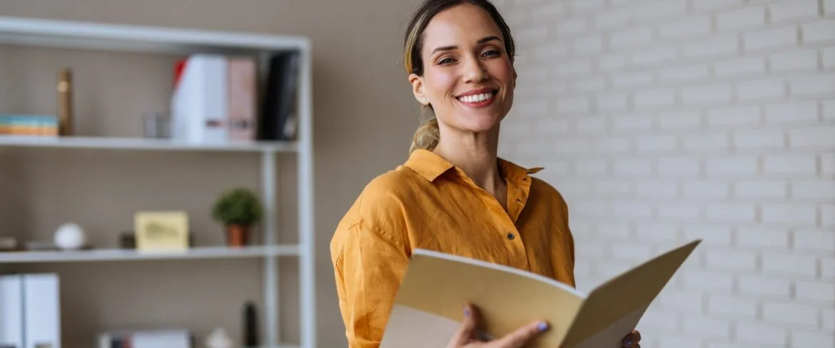 A smiling woman executive assistant in a yellow blouse holds a folder while standing in an office. Behind her, there is a white shelf with books and plants, and a desk with documents and writing tools on a yellow table.