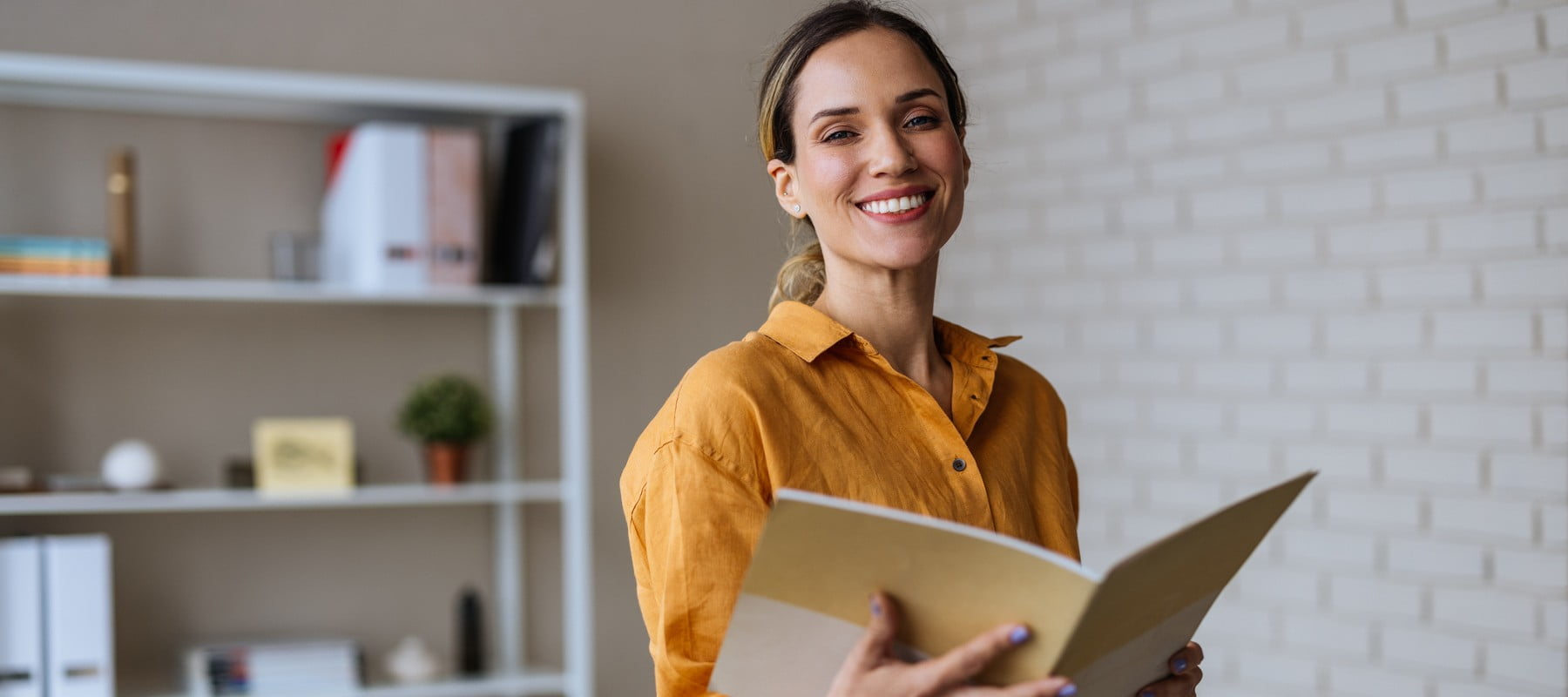 A smiling woman executive assistant in a yellow blouse holds a folder while standing in an office. Behind her, there is a white shelf with books and plants, and a desk with documents and writing tools on a yellow table.