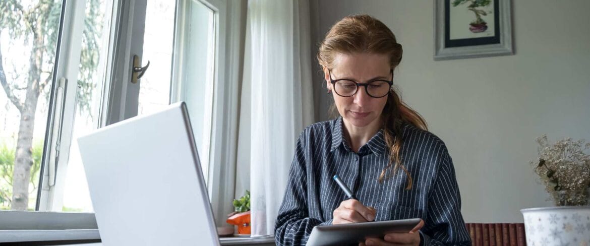 An executive assistant engaged in professional development, wearing glasses and a pinstriped shirt, works at a bright windowsill home office. She focuses on taking notes with a stylus on a tablet while her laptop sits open nearby, demonstrating ongoing skill enhancement. The serene workspace features natural light, houseplants, and a framed botanical print, illustrating an ideal environment for remote professional growth.