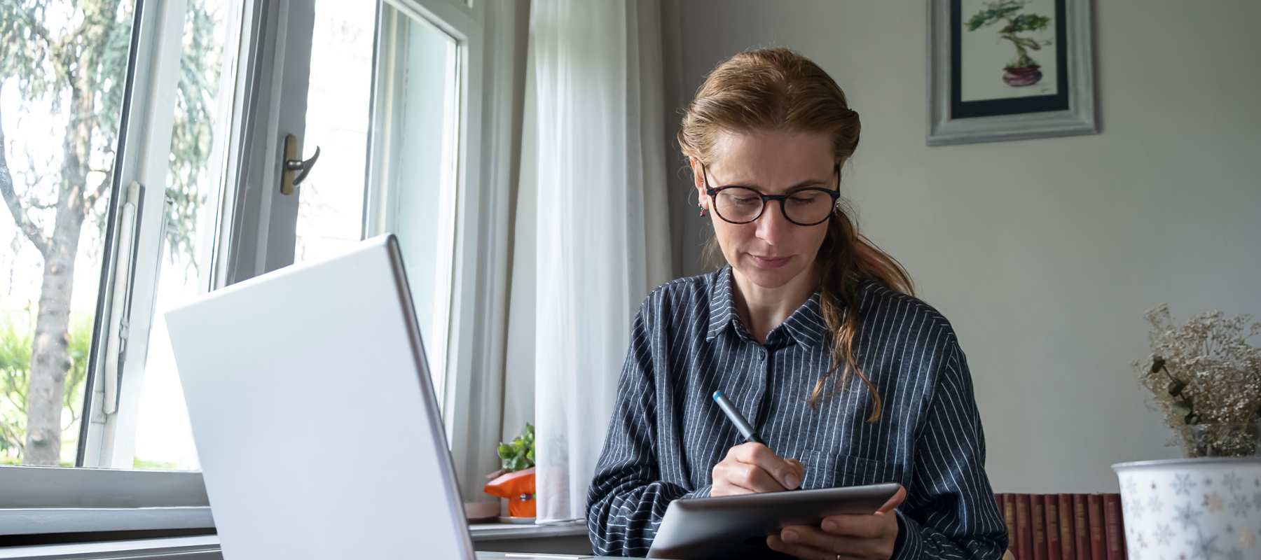 An executive assistant engaged in professional development, wearing glasses and a pinstriped shirt, works at a bright windowsill home office. She focuses on taking notes with a stylus on a tablet while her laptop sits open nearby, demonstrating ongoing skill enhancement. The serene workspace features natural light, houseplants, and a framed botanical print, illustrating an ideal environment for remote professional growth.