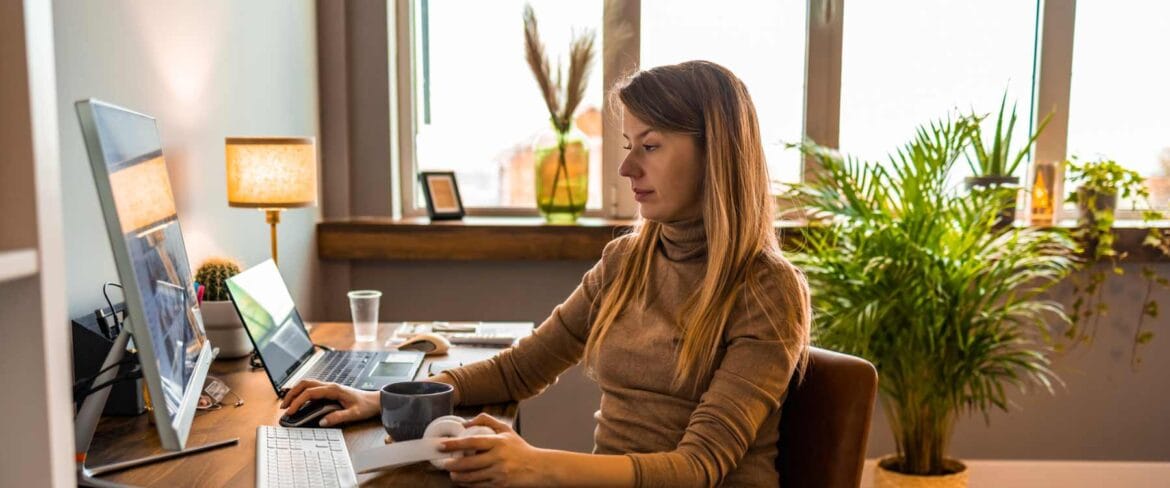 A remote executive assistant works from a modern home office. She is seated at a desk with a large monitor, laptop, keyboard, and mouse. A coffee mug and smartphone are on the desk. The space features natural light, indoor plants, and a cozy atmosphere.