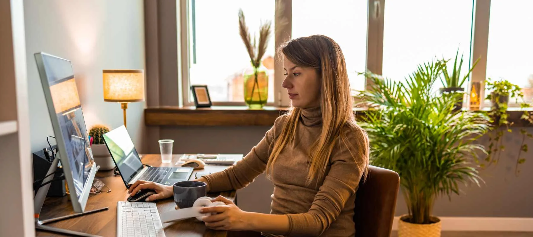 A remote executive assistant works from a modern home office. She is seated at a desk with a large monitor, laptop, keyboard, and mouse. A coffee mug and smartphone are on the desk. The space features natural light, indoor plants, and a cozy atmosphere.