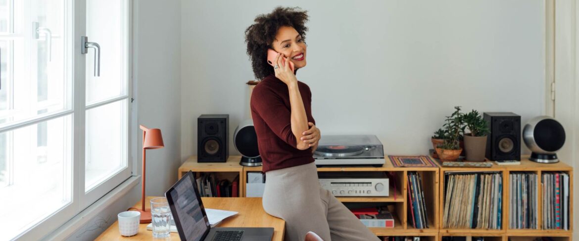 A remote executive assistant smiles while talking on the phone, leaning against a desk in a bright home office with a laptop, records, and bookshelves, illustrating the elevated role of the modern EA and a flexible remote work environment.