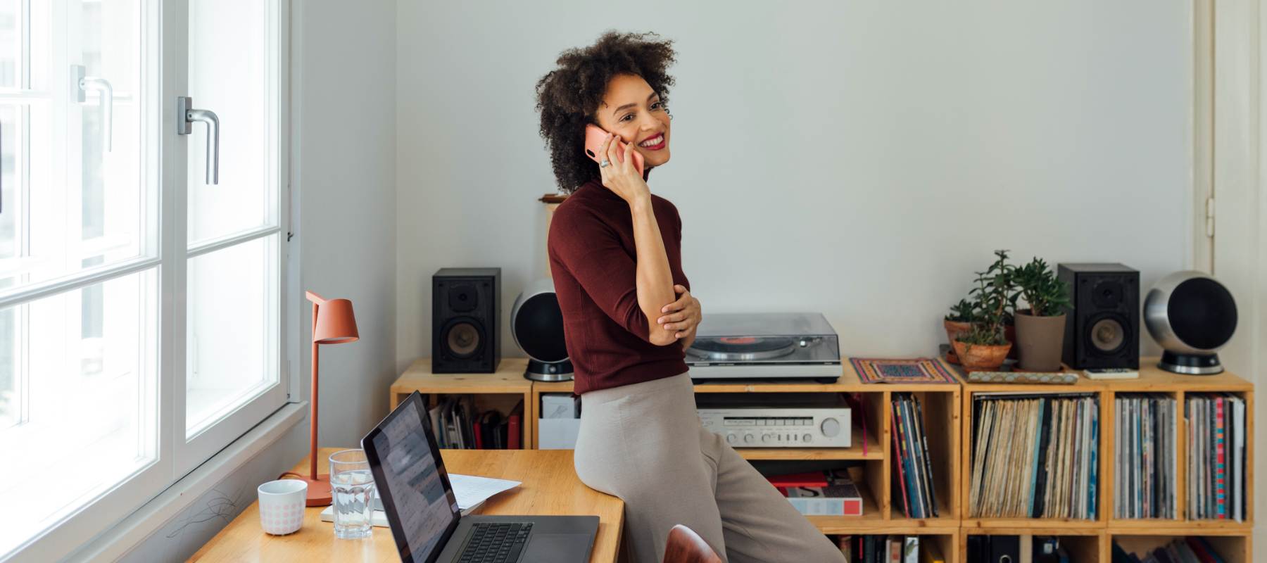 A remote executive assistant smiles while talking on the phone, leaning against a desk in a bright home office with a laptop, records, and bookshelves, illustrating the elevated role of the modern EA and a flexible remote work environment.
