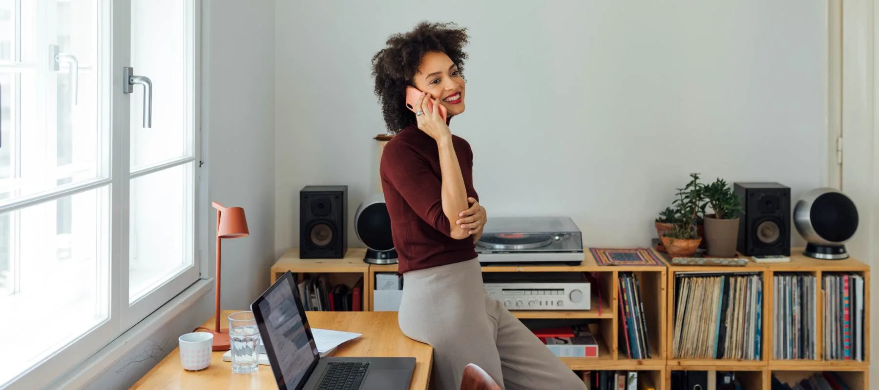 A remote executive assistant smiles while talking on the phone, leaning against a desk in a bright home office with a laptop, records, and bookshelves, illustrating the elevated role of the modern EA and a flexible remote work environment.