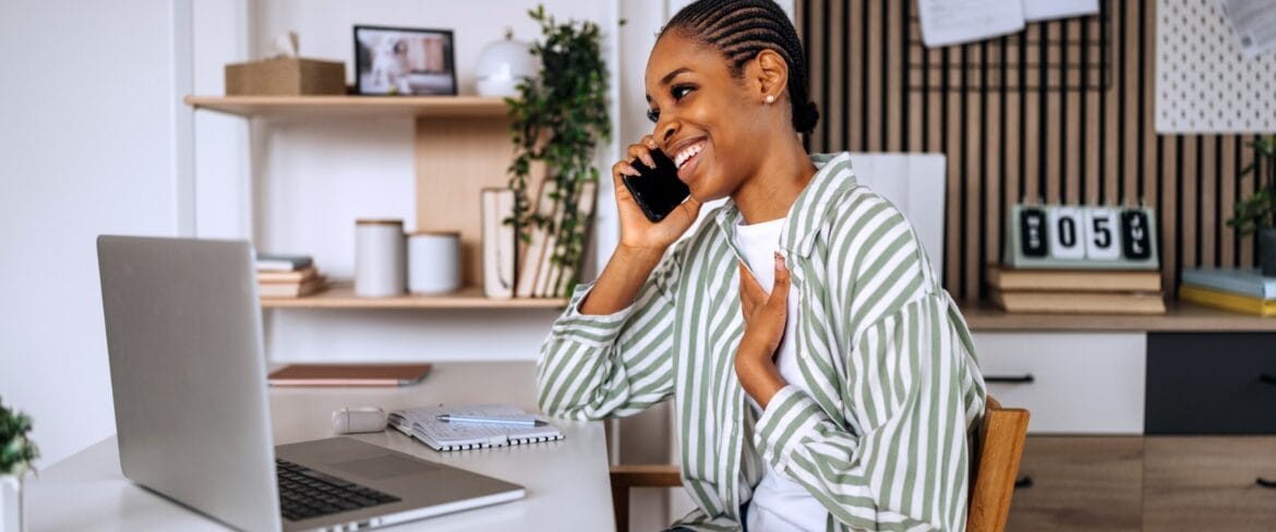 A remote executive assistant smiles during a phone call while working at a tidy home office desk with a laptop, notebook, and calendar, representing the professionalism and communication skills required to support high-level executives virtually.