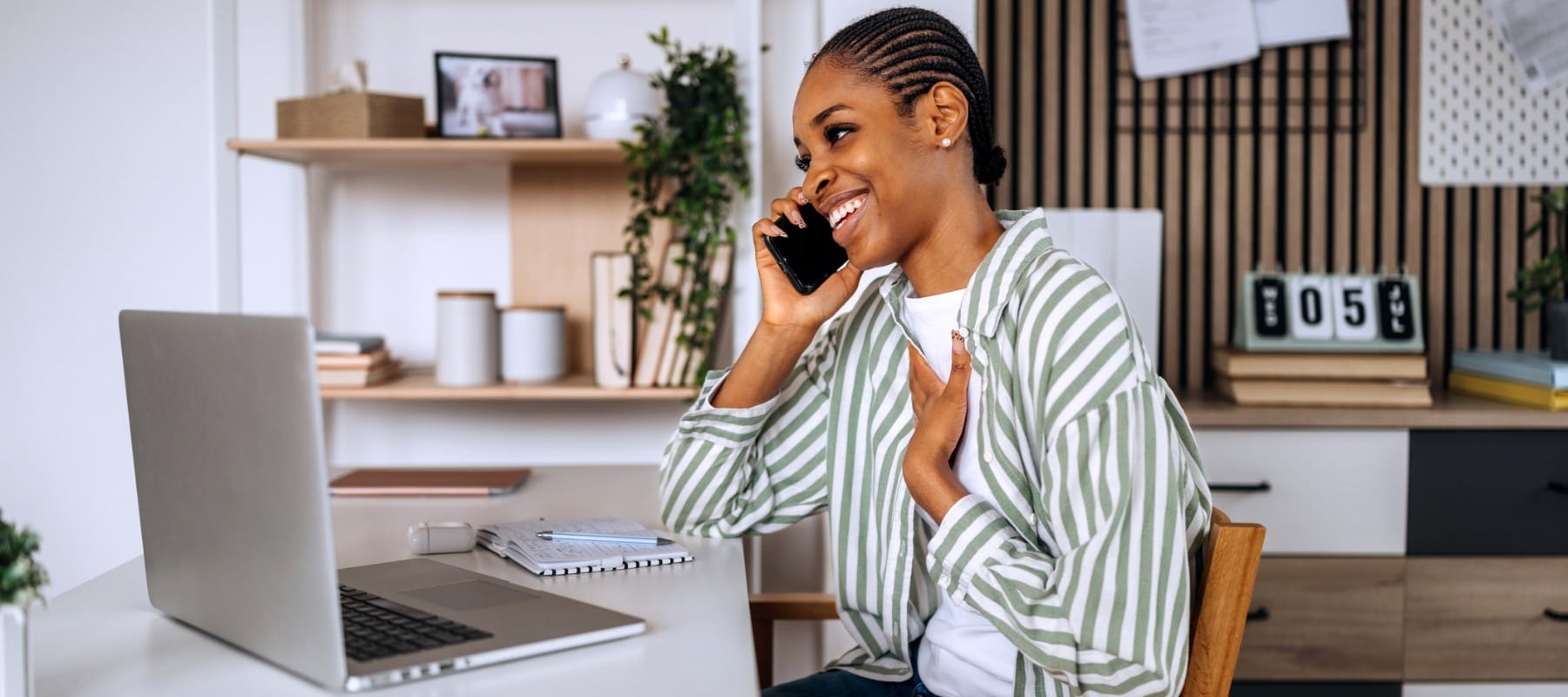 A remote executive assistant smiles during a phone call while working at a tidy home office desk with a laptop, notebook, and calendar, representing the professionalism and communication skills required to support high-level executives virtually.