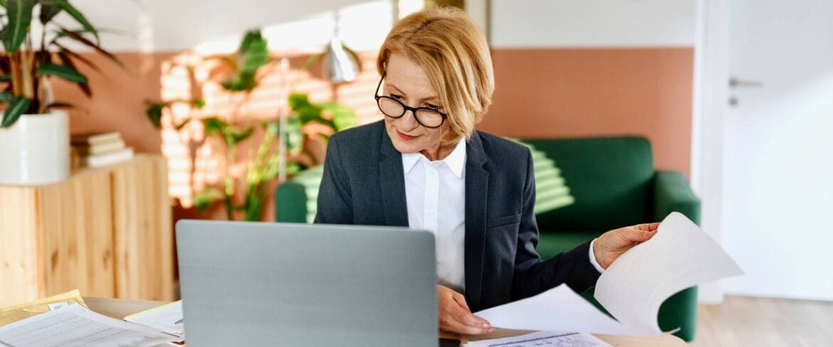 Professional woman wearing glasses and business suit reviewing documents at laptop in modern office with plants and green couch, representing the administrative tasks of a remote executive assistant.