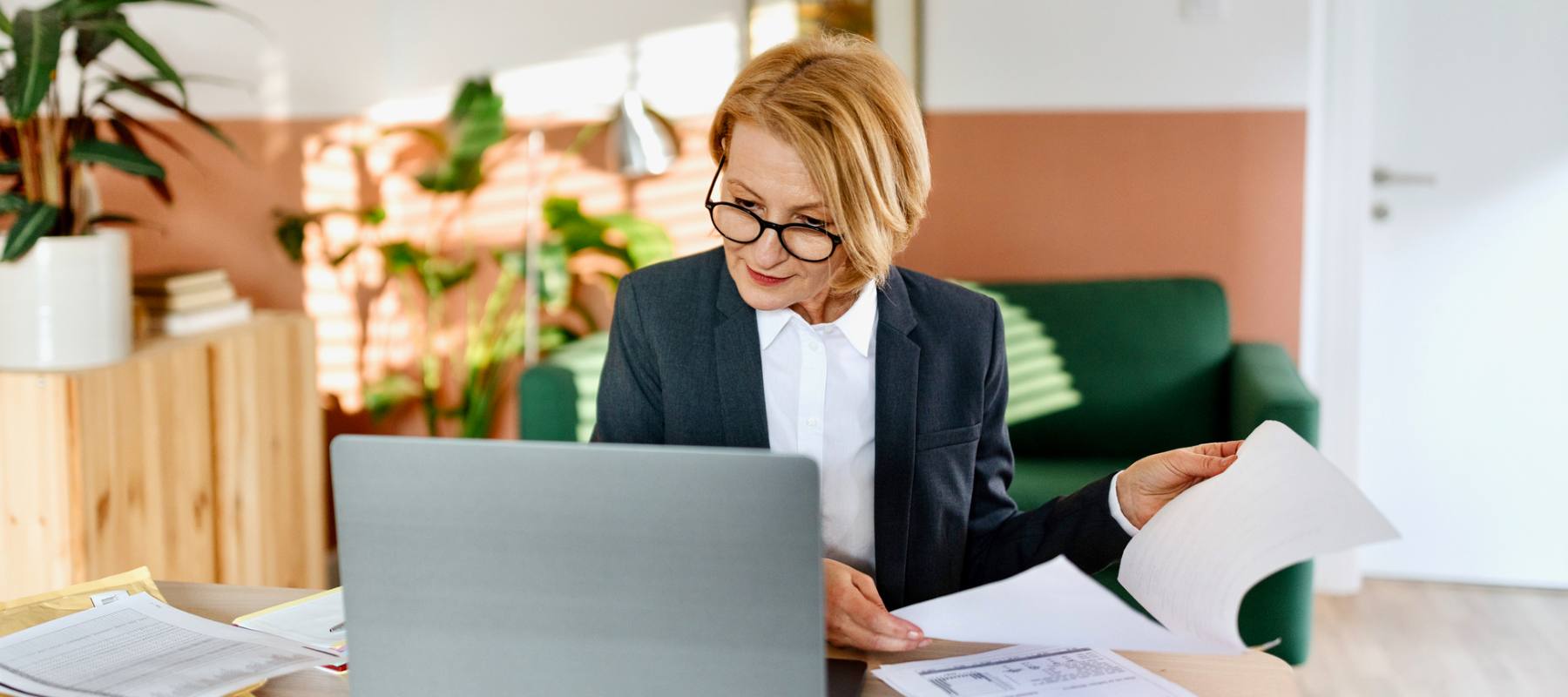 Professional woman wearing glasses and business suit reviewing documents at laptop in modern office with plants and green couch, representing the administrative tasks of a remote executive assistant.
