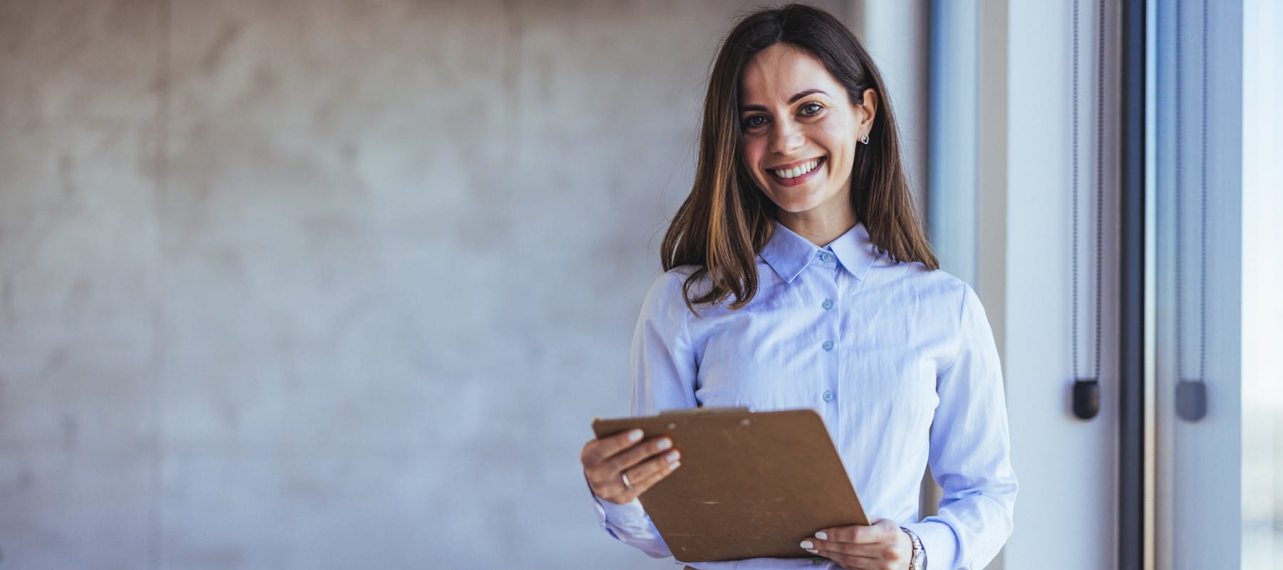Professional woman in a light blue button-down shirt smiling confidently while holding a clipboard in a modern office setting.