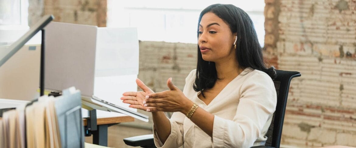 Remote executive assistant in business attire gesturing while speaking during a video call at her desk in a modern office with exposed brick walls, illustrating the challenges of remote work communication and hiring.