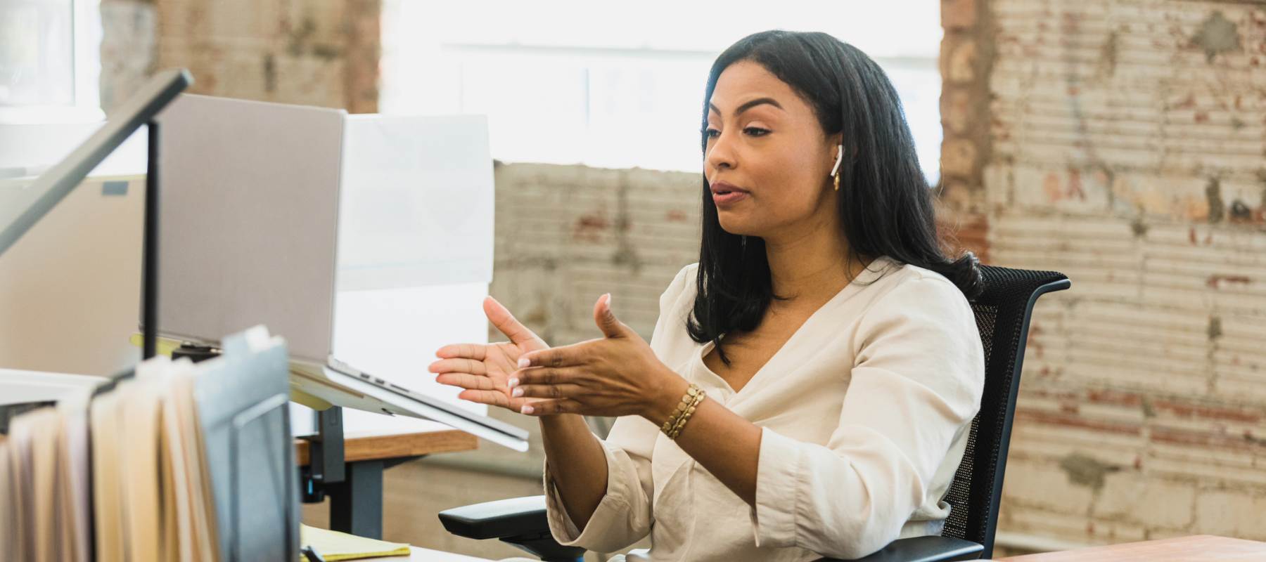Remote executive assistant in business attire gesturing while speaking during a video call at her desk in a modern office with exposed brick walls, illustrating the challenges of remote work communication and hiring.