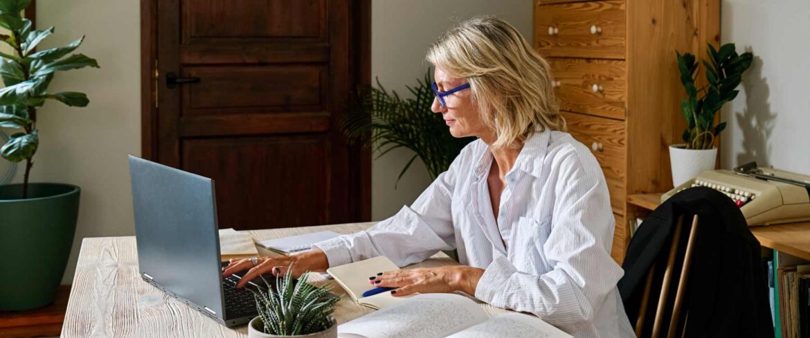 An executive assistant works from a private home office, reviewing documents and typing on a laptop at a wooden desk. The quiet, organized setting and focused posture reflect the confidentiality and discretion required when handling sensitive information.