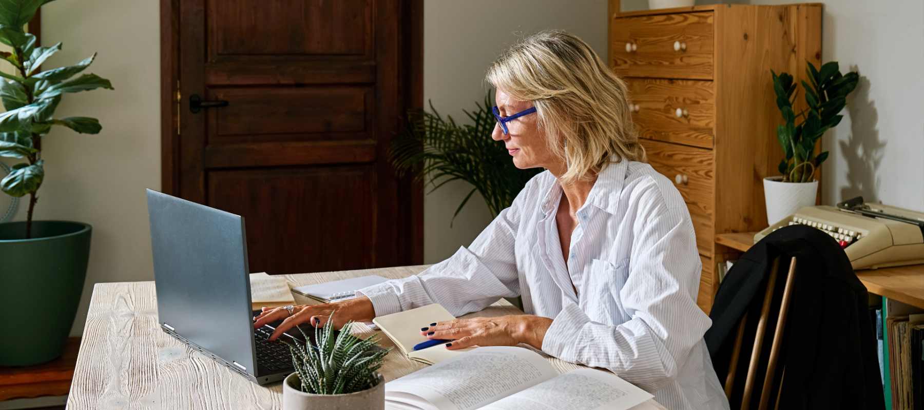 An executive assistant works from a private home office, reviewing documents and typing on a laptop at a wooden desk. The quiet, organized setting and focused posture reflect the confidentiality and discretion required when handling sensitive information.