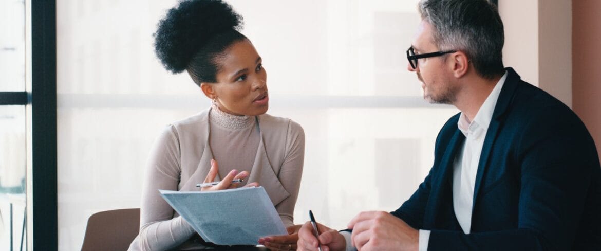 An executive assistant meets with a business leader in a professional office setting, reviewing documents and discussing details face-to-face, highlighting the collaborative and communication-focused nature of high-level administrative support.