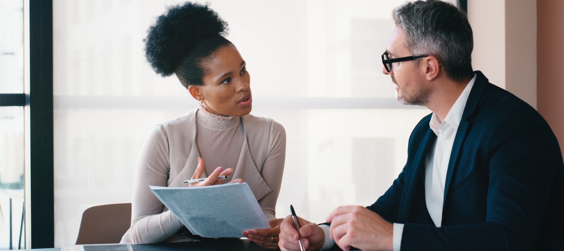 An executive assistant meets with a business leader in a professional office setting, reviewing documents and discussing details face-to-face, highlighting the collaborative and communication-focused nature of high-level administrative support.
