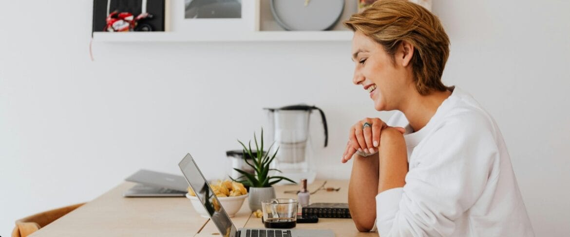 Smiling executive assistant working remotely from a bright home office, seated at a desk with a laptop, coffee, and workspace essentials. The image accompanies guidance on how to find a qualified executive assistant replacement during a leave of absence or role transition.