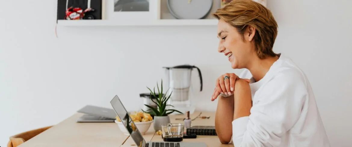 Smiling executive assistant working remotely from a bright home office, seated at a desk with a laptop, coffee, and workspace essentials. The image accompanies guidance on how to find a qualified executive assistant replacement during a leave of absence or role transition.